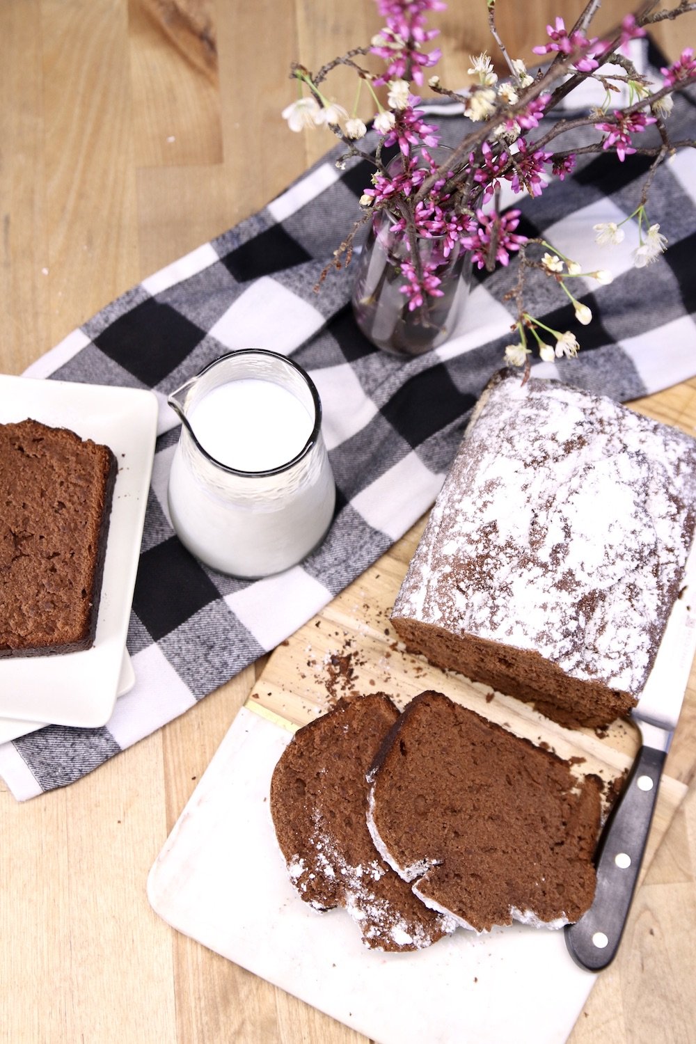 Sliced banana bread with loaf, jar of  milk, vase of red bud stems, black and white napkin