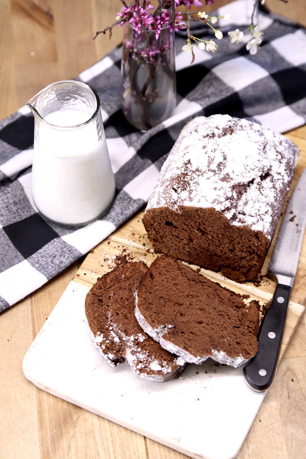 Chocolate banana bread with a few slices off of the loaf, milk and flowers on the table