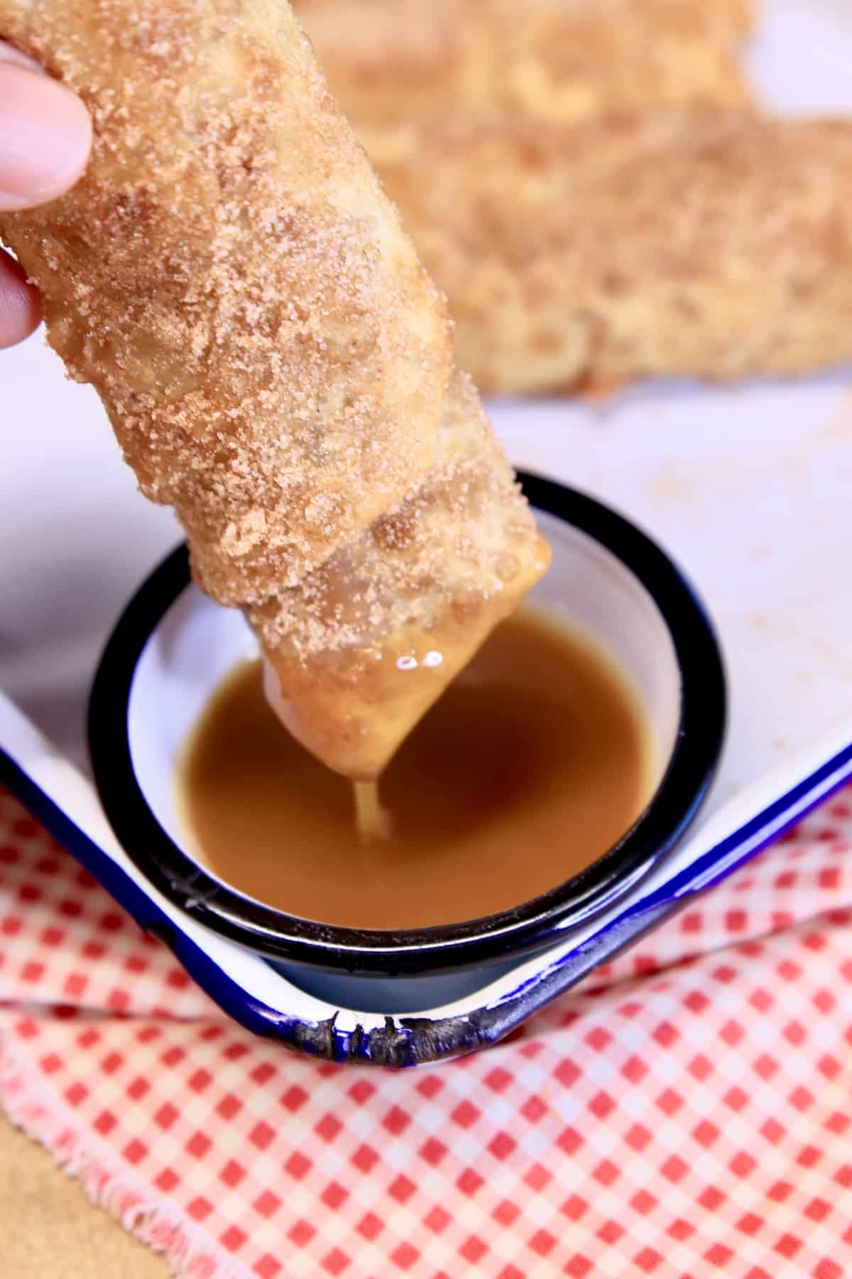 Apple pie egg roll dipping into bowl of caramel sauce.