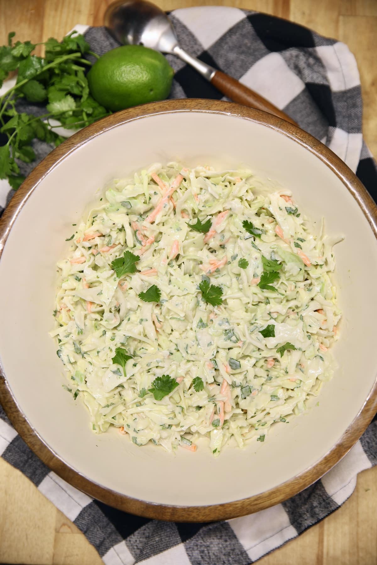 Bowl of cilantro lime slaw on a checked napkin, cilantro and lime beside bowl.