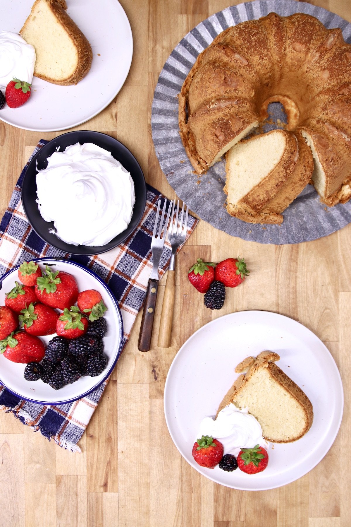 overhead photo of cake on cake plate, 2 slices on saucers with whipped cream & berries, bowls of whipped cream and berries