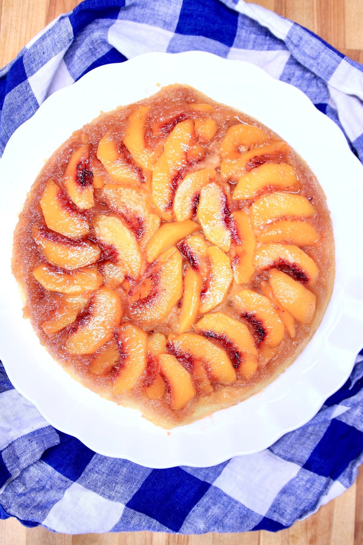 Peach upside down cake on a white platter, blue and white check cloth beneath.