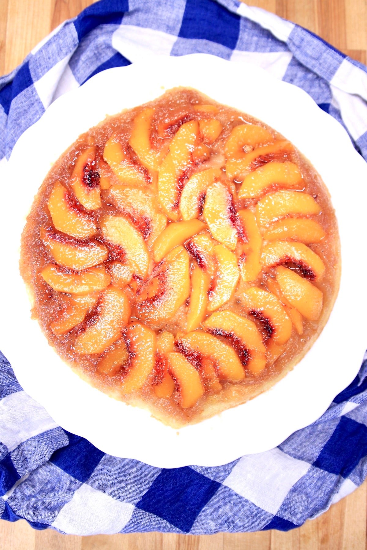 Overhead view of peach upside down cake on a white platter, blue and white check napkin.