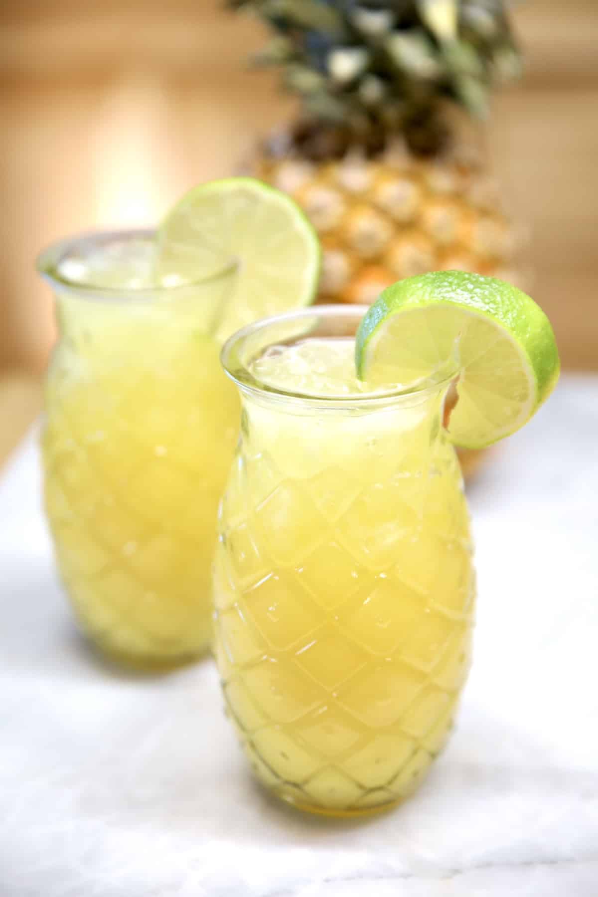 2 tiki glasses with pineapple margaritas, pineapple in background.