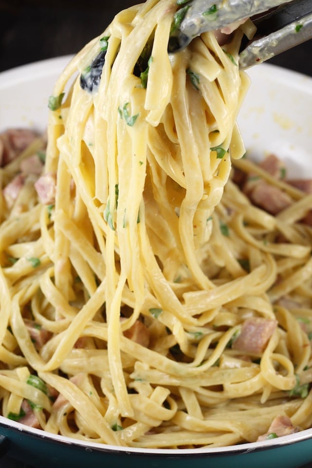 Tongs grabbing pasta and ham from skillet