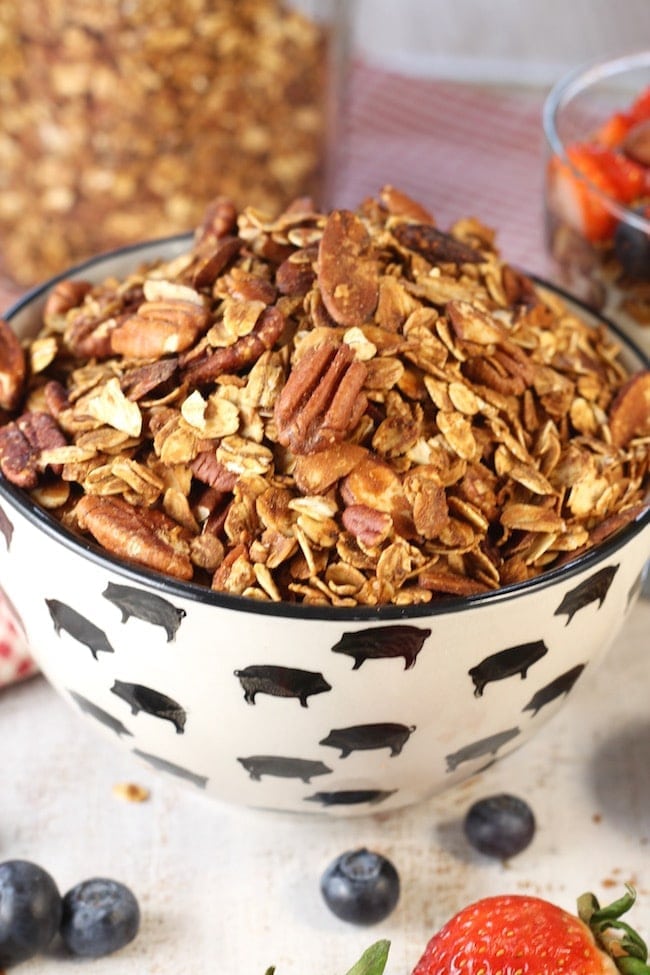 Easy granola recipe with pecans and almonds in a black and white bowl with blueberries and strawberries on the white back ground