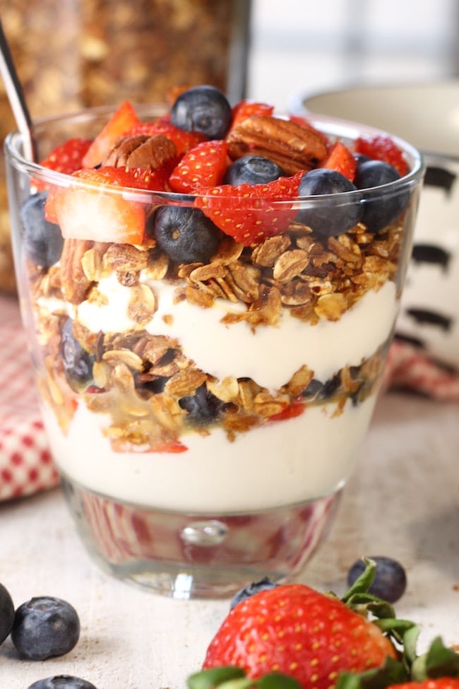 Parfait Glass filled with vanilla yogurt, easy homemade granola, strawberries and blueberries on a white cutting board with red check napkin and fresh berries