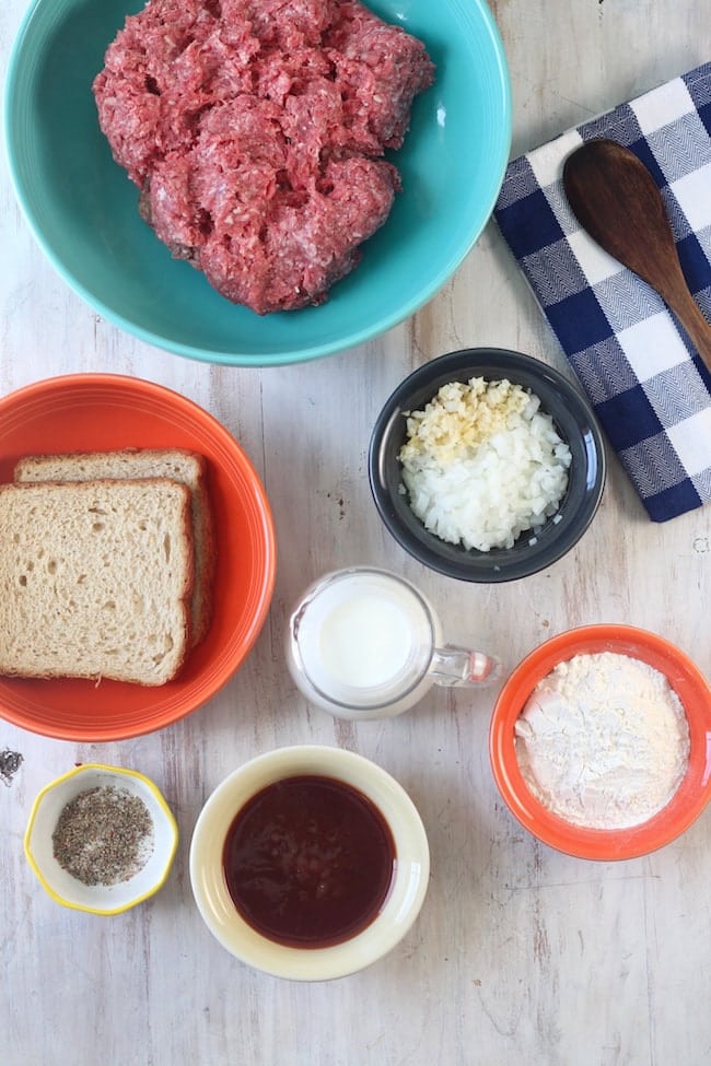 2 slices of wheat bread, ground beef in a blue bowl, chopped onions and garlic in a bowl, milk in a clear pitcher, flour in a bowl, barbecue sauce in a bowl, salt and pepper in a bowl, blue check napkin with a wood spoon laying on it ~ Ingredients for Barbecue Meatballs
