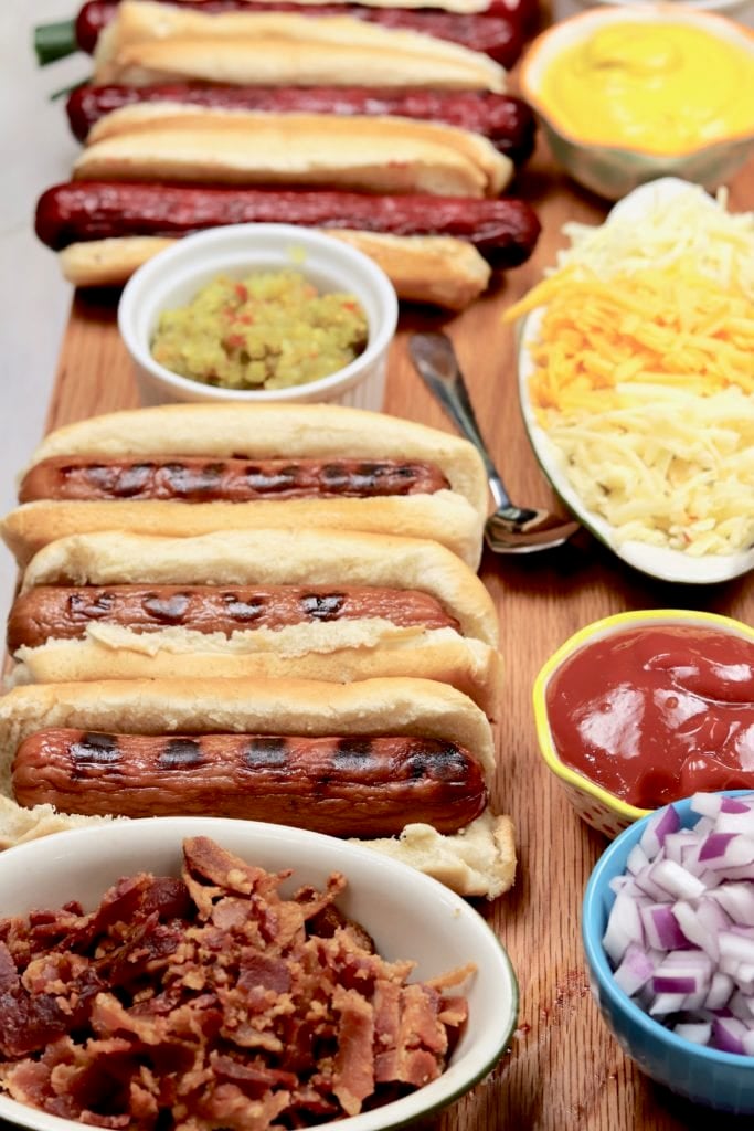 Hot Dogs on a board with bacon, onion, ketchup and shredded cheese