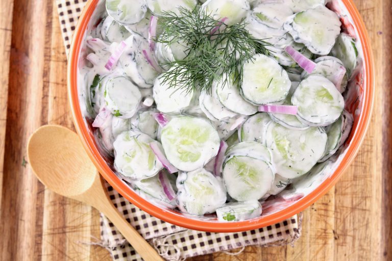Bowl of sliced cucumbers, red onion and dill in a creamy sauce on a wood board with bamboo spoon