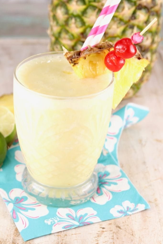 Frozen pineapple mocktail garnished with maraschino cherries, fresh pineapple and a pink stiped straw on a blue napkin