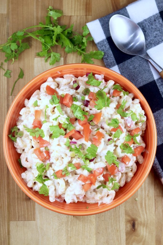 Pasta salad in an orange bowl with tomatoes, cilantro and onions. Black and white check napkin and fresh cilantro in background