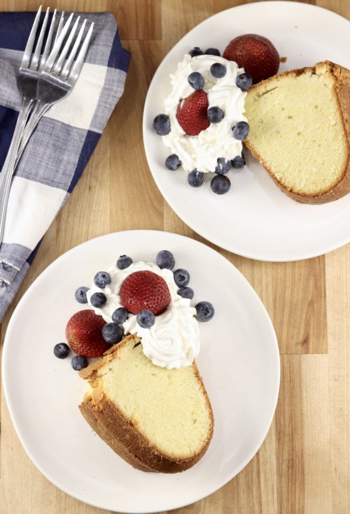 Slices of pound cake on 2 plates with whipped cream, strawberries and blueberries, blue napkin