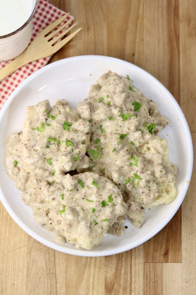 Sausage gravy and biscuits on a plate with glass of milk and bamboo fork to the side