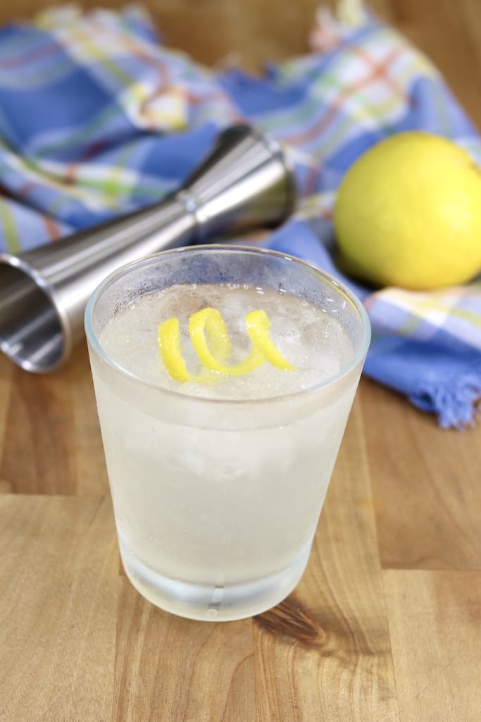 Gin Sour Cocktail in a old fashioned glass with a lemon twist - blue napkin and jigger in the background with a whole lemon