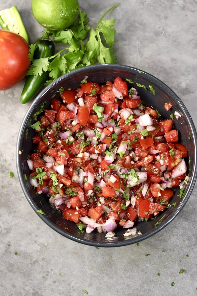 Pico de gallo salsa in a black bowl with cilantro, and tomato by the bowl. Gray background, overhead view.