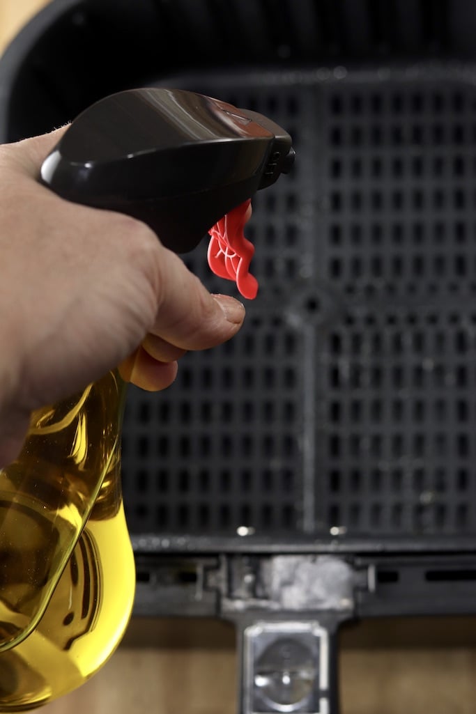 spraying air fryer basket with olive oil in a spray bottle