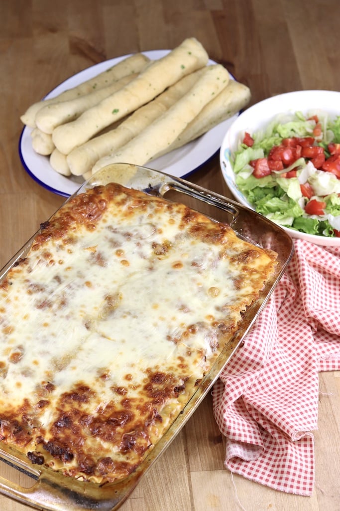 Lasagna in a casserole dish, plate of breadsticks, salad in a bowl with tomatoes, red check napkin