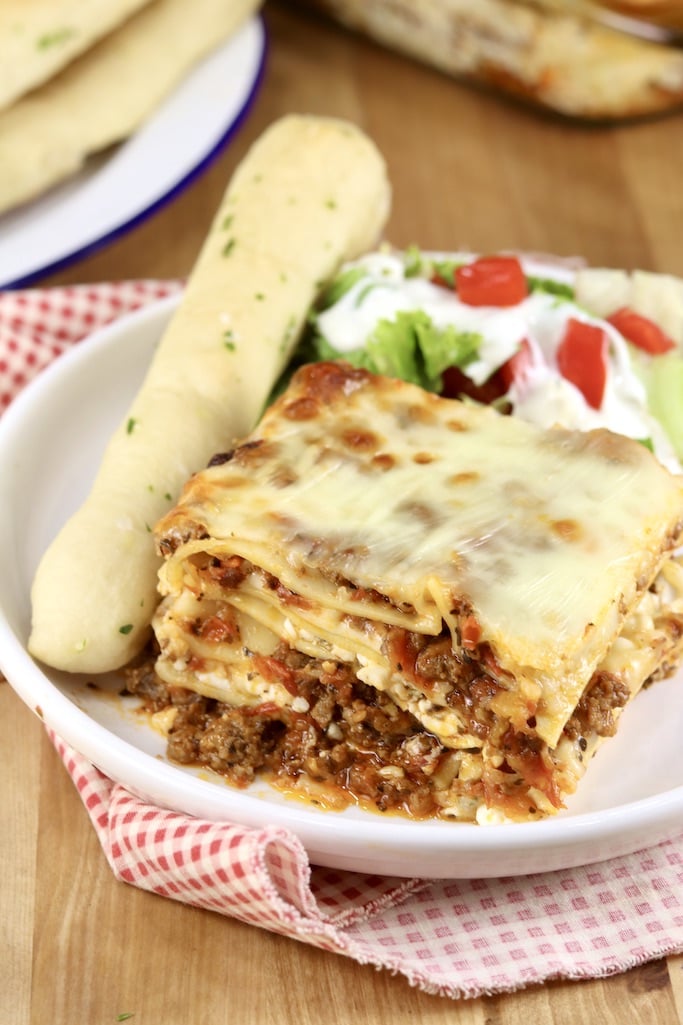 Slice of baked lasagna with a breadstick and salad on a white plate