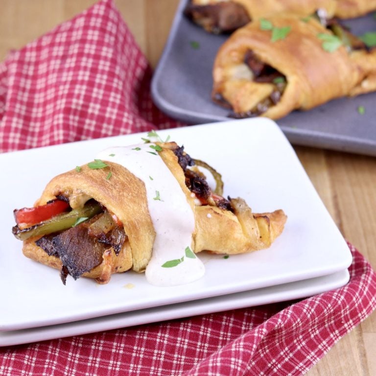 Cheesesteak Crescent Roll Ups with horseradish sauce on a white plate, platter of rolls in back ground. Red check napkin