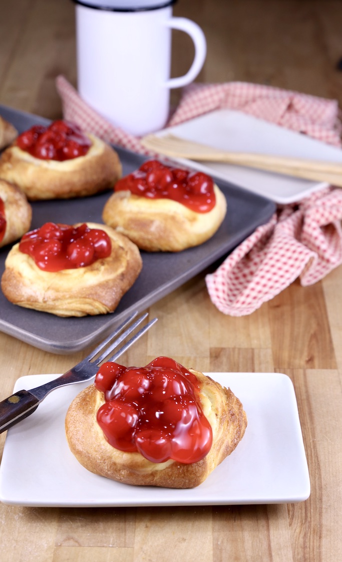 Saucer with cherry cheese danish and a fork, platter of pastries are in background