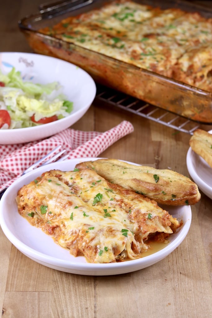 Plated manicotti with a breadstick, salad and pan of manicotti in background