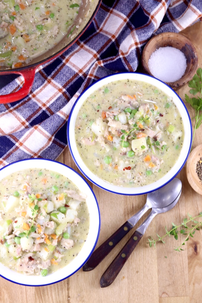 2 bowls of creamy chicken soup, edge of soup pan and blue napkin. 2 spoons, bowl of salt and herbs