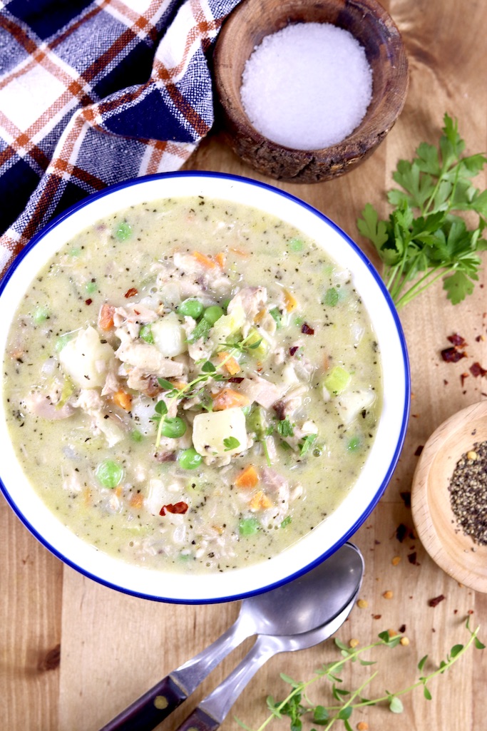 Chicken Pot Pie Soup in a white bowl, salt, herbs and spoons with a plaid napkin surround the bowl