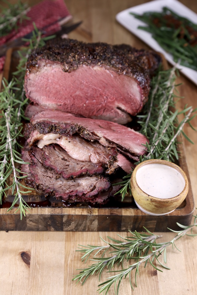 Overhead view of sliced prime rib roast with horseradish sauce, fresh rosemary, platter of green beans in background