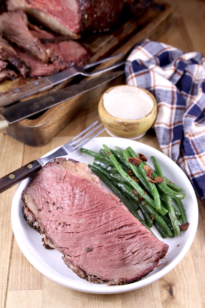 Slice of prime rib on a plate with green beans, horseradish sauce, plaid napkin