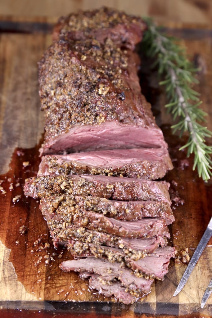 Beef tenderloin on a cutting board, half of the roast sliced. Sprig of rosemary to the side