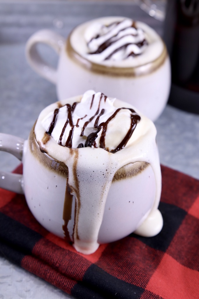 2 mugs of mudslide coffee with whipped cream and a drizzle of chocolate, overflowing onto a red check napkin