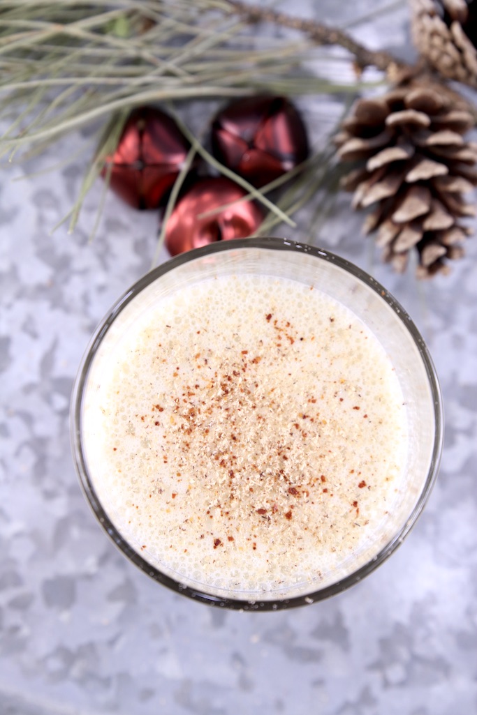 Eggnog cocktail garnished with nutmeg and red pepper on a galvanized tray with pinecone, greenery and 3 red jingle bells