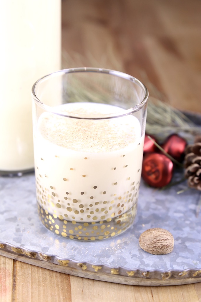 Spiked Eggnog in an old fashioned glass on a tray with holiday ornaments, and whole nutmeg. Tall pitcher of eggnog in background