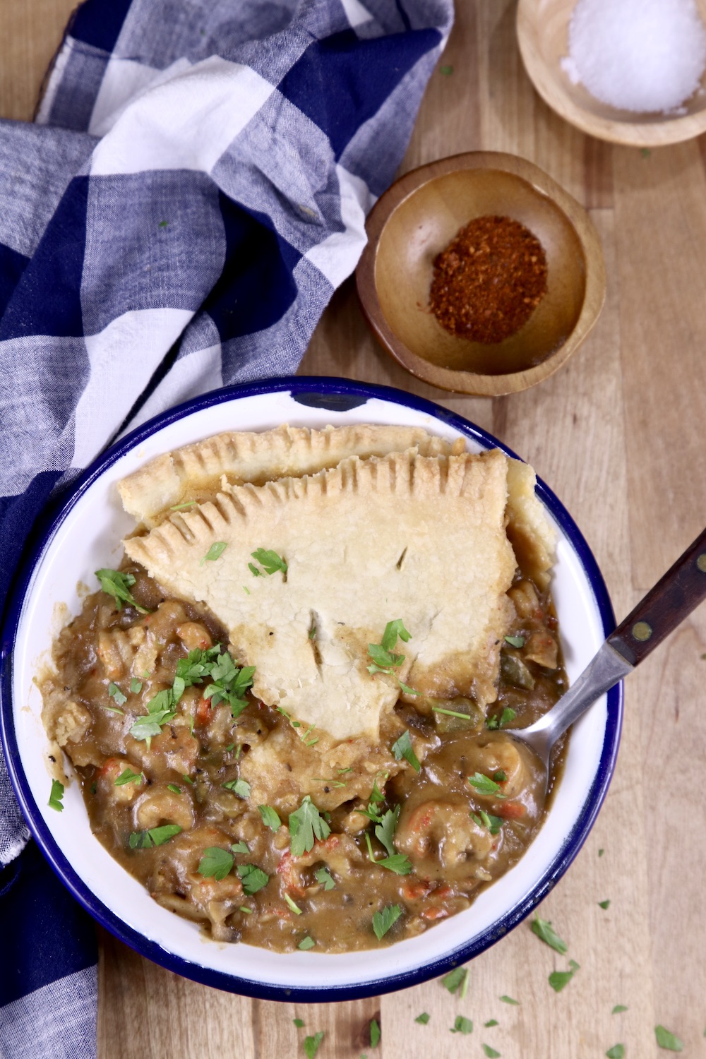 Overhead view of crawfish pie served in a blue rimmed bowl. Blue check napkin, small bowl of red pepper, small bowl of salt