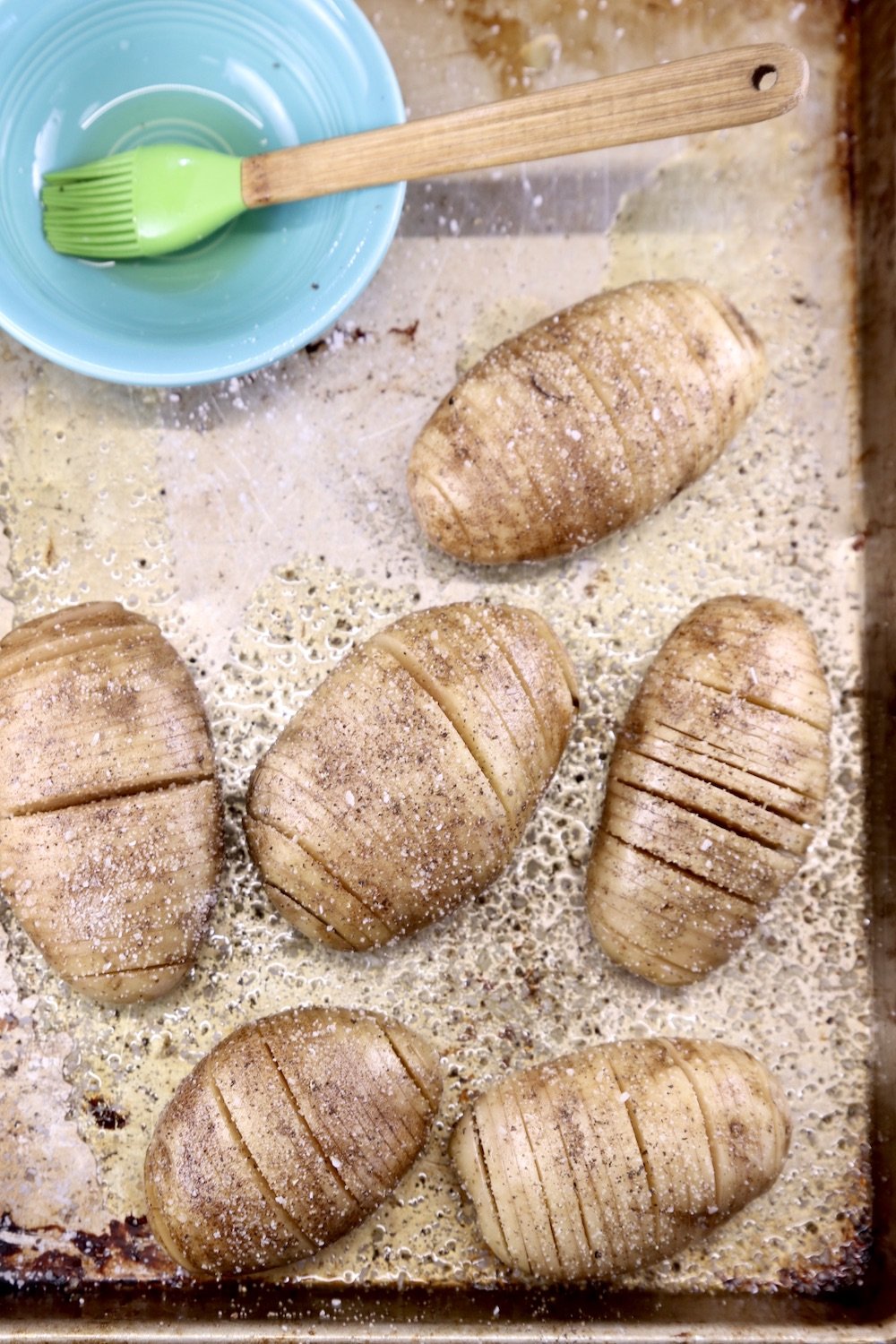 Sheet pan of fan sliced potatoes brushed with olive oil, bowl of oil and brush to the side