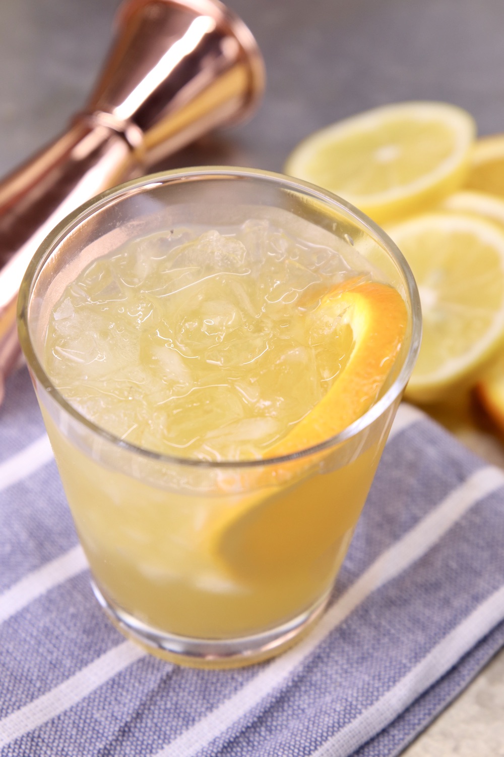 Amaretto Whiskey Sour garnished with orange slice on a blue and white stripe towel with a copper jigger and lemon slices in background