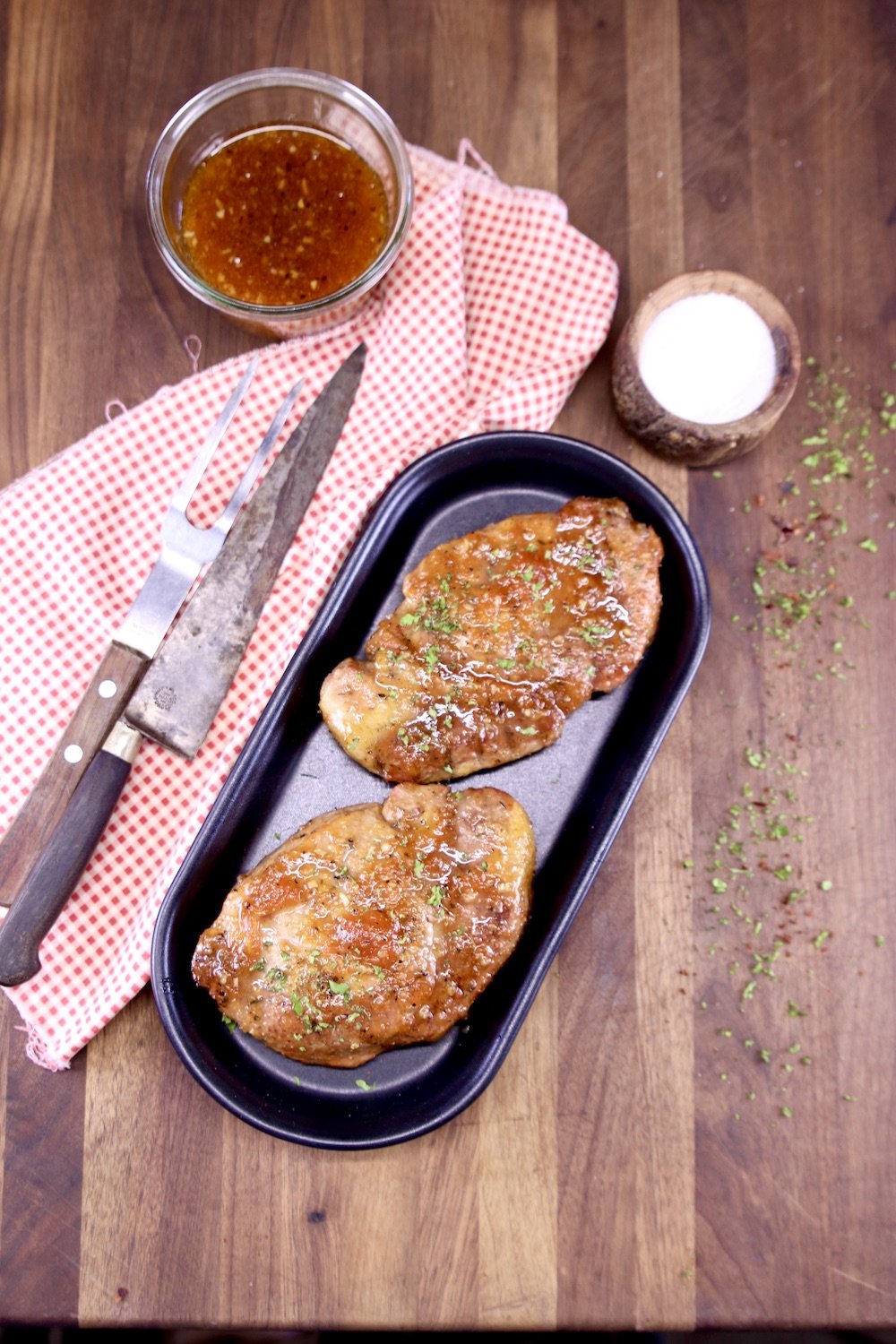 2 grilled pork chops with jar of sauce, bowl of salt, knife and serving fork on a red napkin