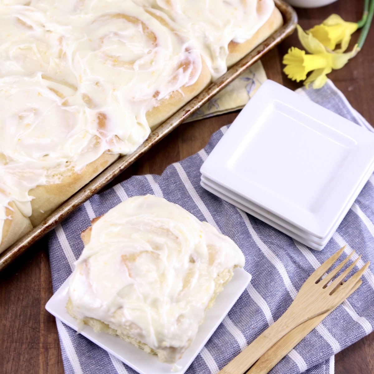 Pineapple sweet rolls with cream cheese icing. One on a small dessert plate, tray of rolls in corner