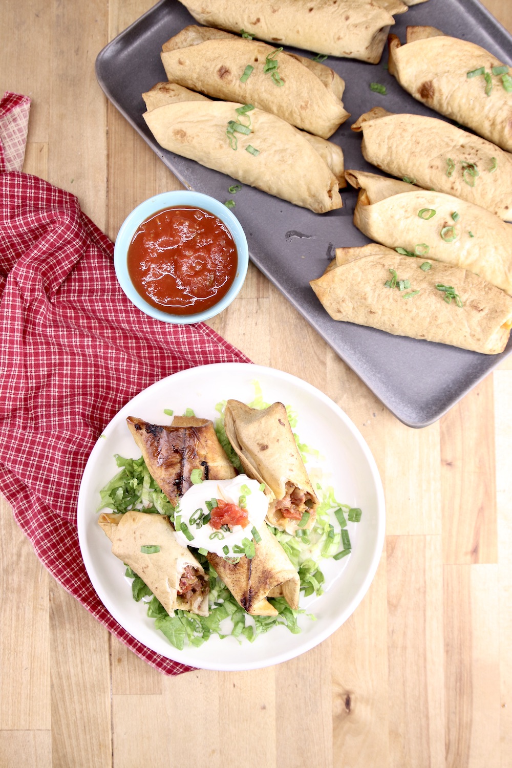overhead view of burritos, plated with lettuce, sour cream & salsa. Platter of burritos and small bowl of salsa.