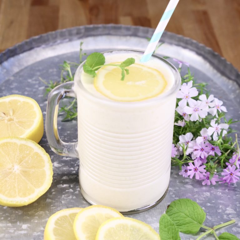 frosted pineapple lemonade in a glass with straw and slice of lemon. Sitting on a tray with lemon slices and small flowers