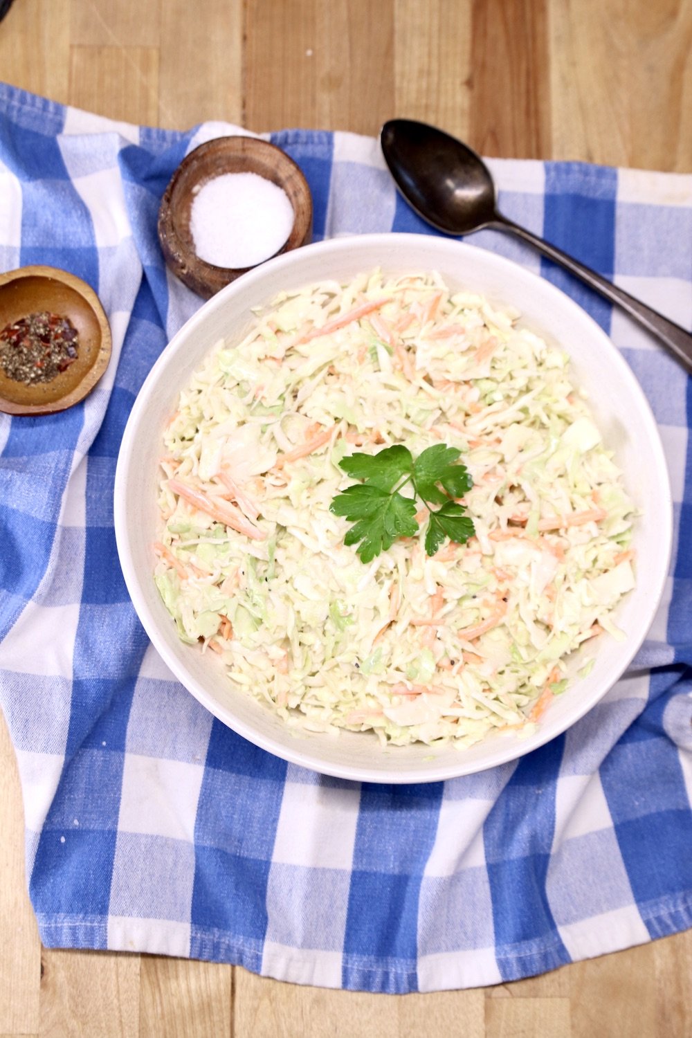 coleslaw in a bowl on a blue napkin, spoon, small bowls of salt & pepper all on a blue check napkin
