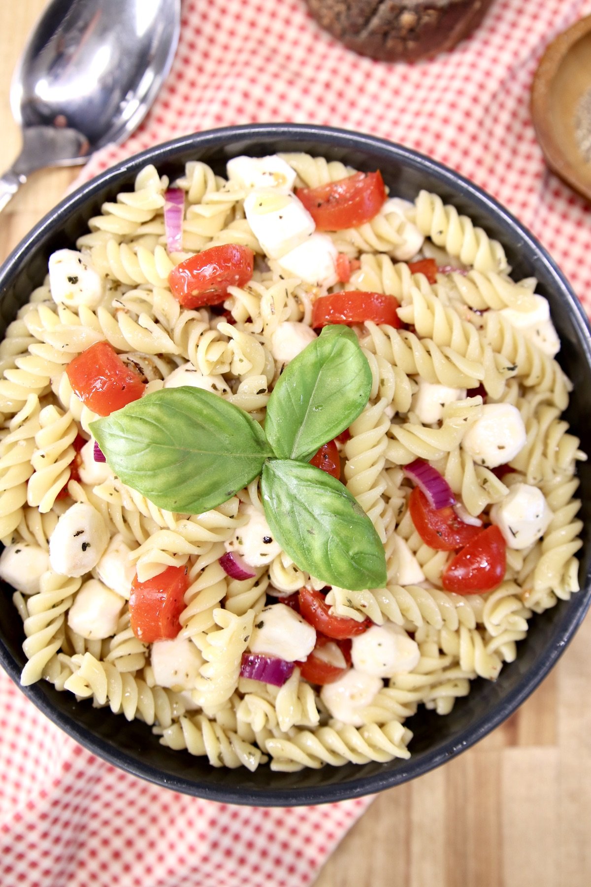 overhead view of pasta salad in a black bowl. Tomatoes, mozzarella and basil garnish