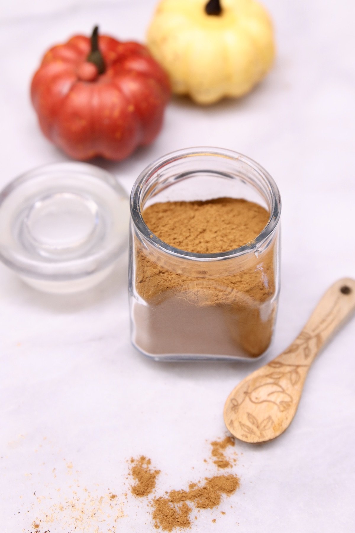 jar of pumpkin pie spice with 2 decorative pumpkins and wood spoon