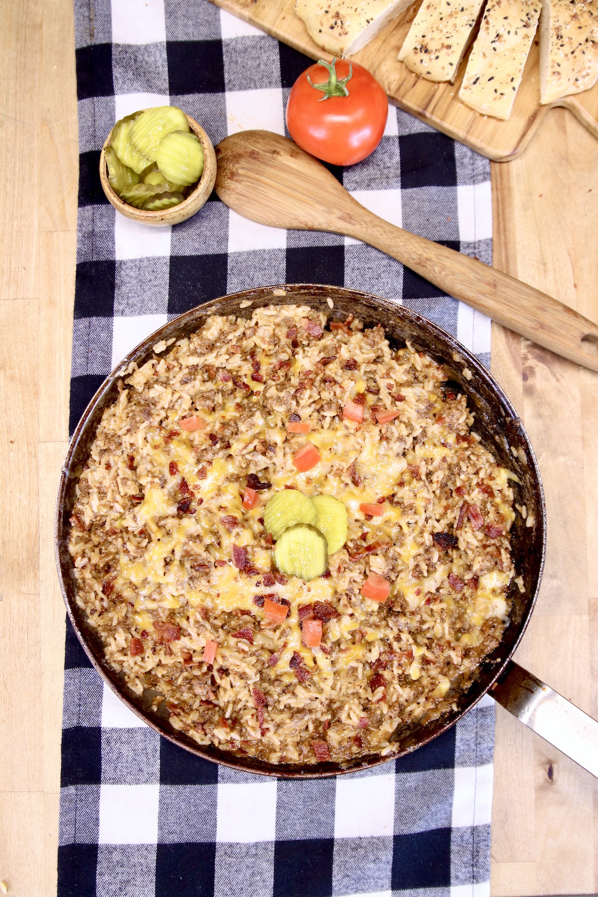 overhead bacon cheeseburger skillet on a black and white check runner. Wooden spoon, pickles, tomato.