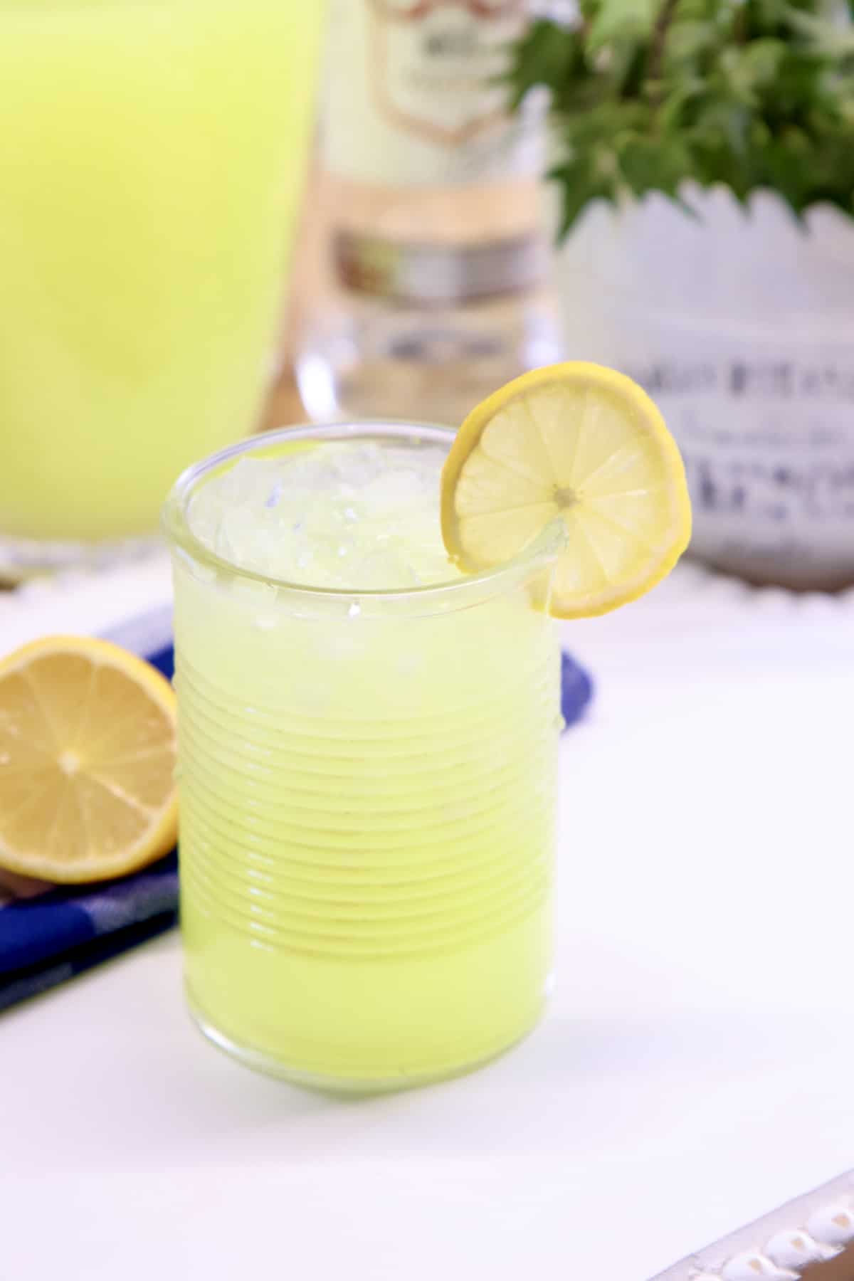 vodka lemonade cocktail in a glass with lemon garnish. Ivy plant and pitcher of lemonade in background.