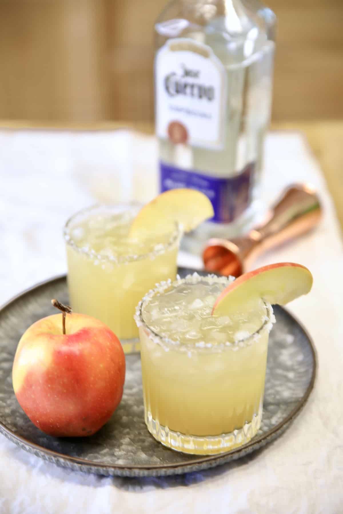 2 margaritas on a tray with apple. Tequila in background.