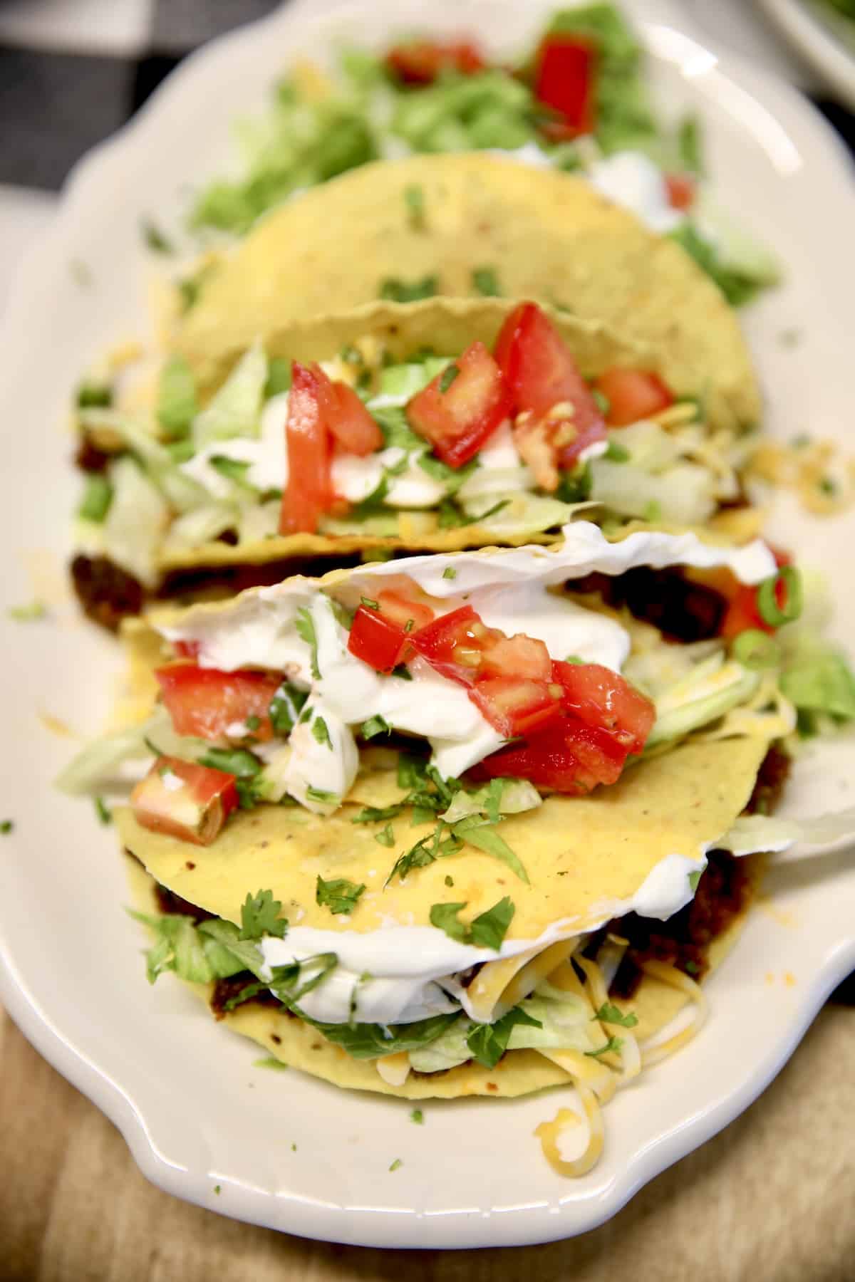 Ground beef tacos on a platter with tomatoes, lettuce, cheese, sour cream.