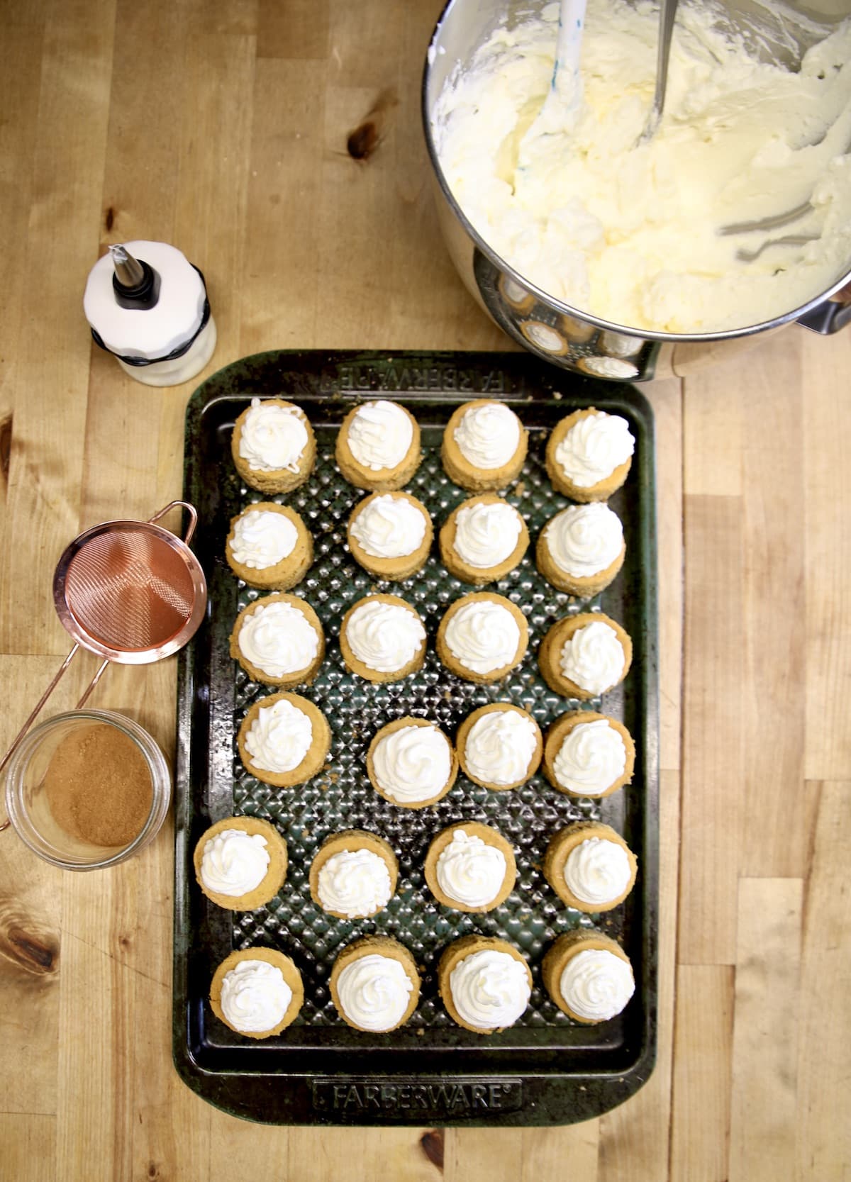 Overhead view of mini pumpkin cheesecakes on a pan, bowl of whipped cream, pumpkin pie spice.