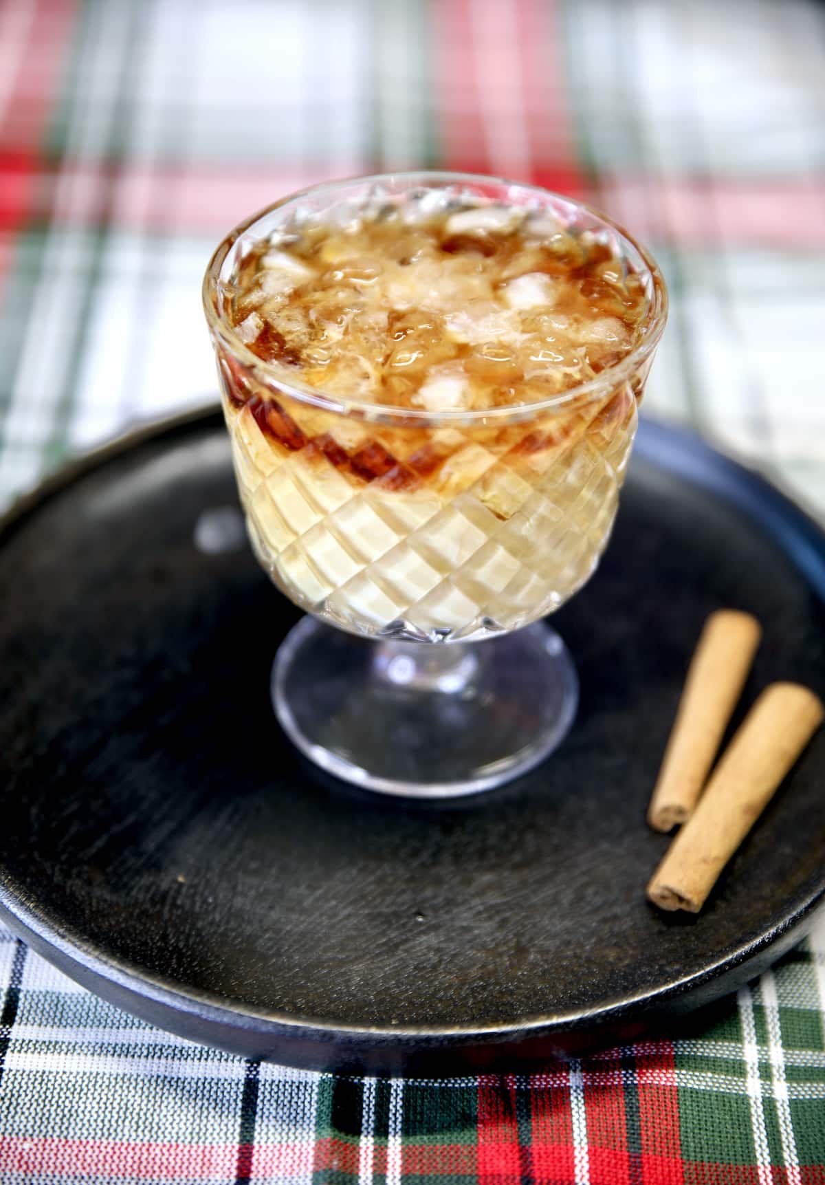 White Russian cocktail on a black plate, 2 cinnamon sticks. On Christmas plaid cloth.